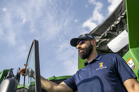 A bearded man in a hat stands next to large farming equipment