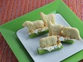 4 pieces of celery with filling and topped with different animal crackers. The snack is place on a small white square plate, with a larger green square place underneath. All of this is on a stripped red and green background.