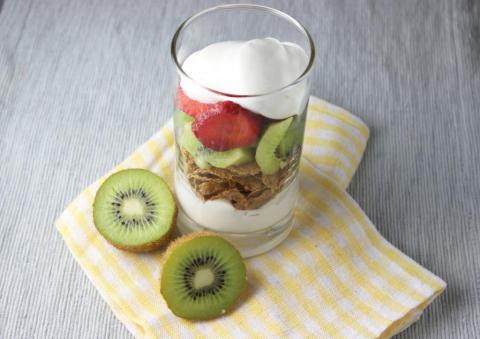 On a gray background sits a drinking glass filled with layers of kiwi fruit, strawberries, cereal, and yogurt. A kiwi fruit is cut in half, with both halves arranged near the parfait. Both parfait and kiwi fruit halves sit on a folded yellow and white napkin.