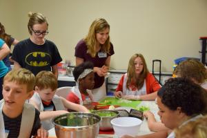 female staff member standing near a group of youth at a table preparing food to make a recipe