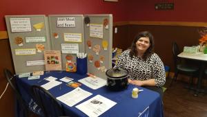 staff member sitting at a booth with nutrition education materials and a trifold board about lean meats and beans