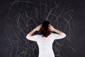 Woman with hands on head facing a blackboard full of chaotic chalk marks