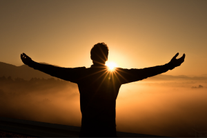 Man with widespread, open arms facing a bright sunrise