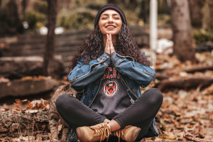 Happy young woman sitting cross-legged on forest floor with hands together in meditation