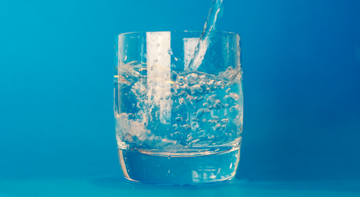 Blue background, glass in the middle of picture with water being poured into it.