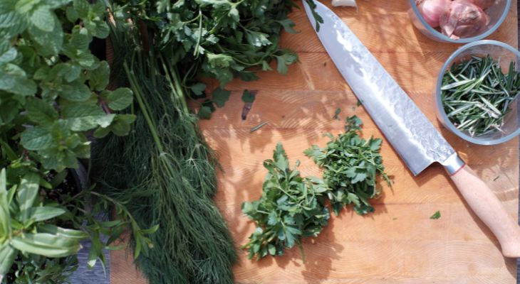 a mix of green herbs sit on a chopping block next to a large kitchen knife
