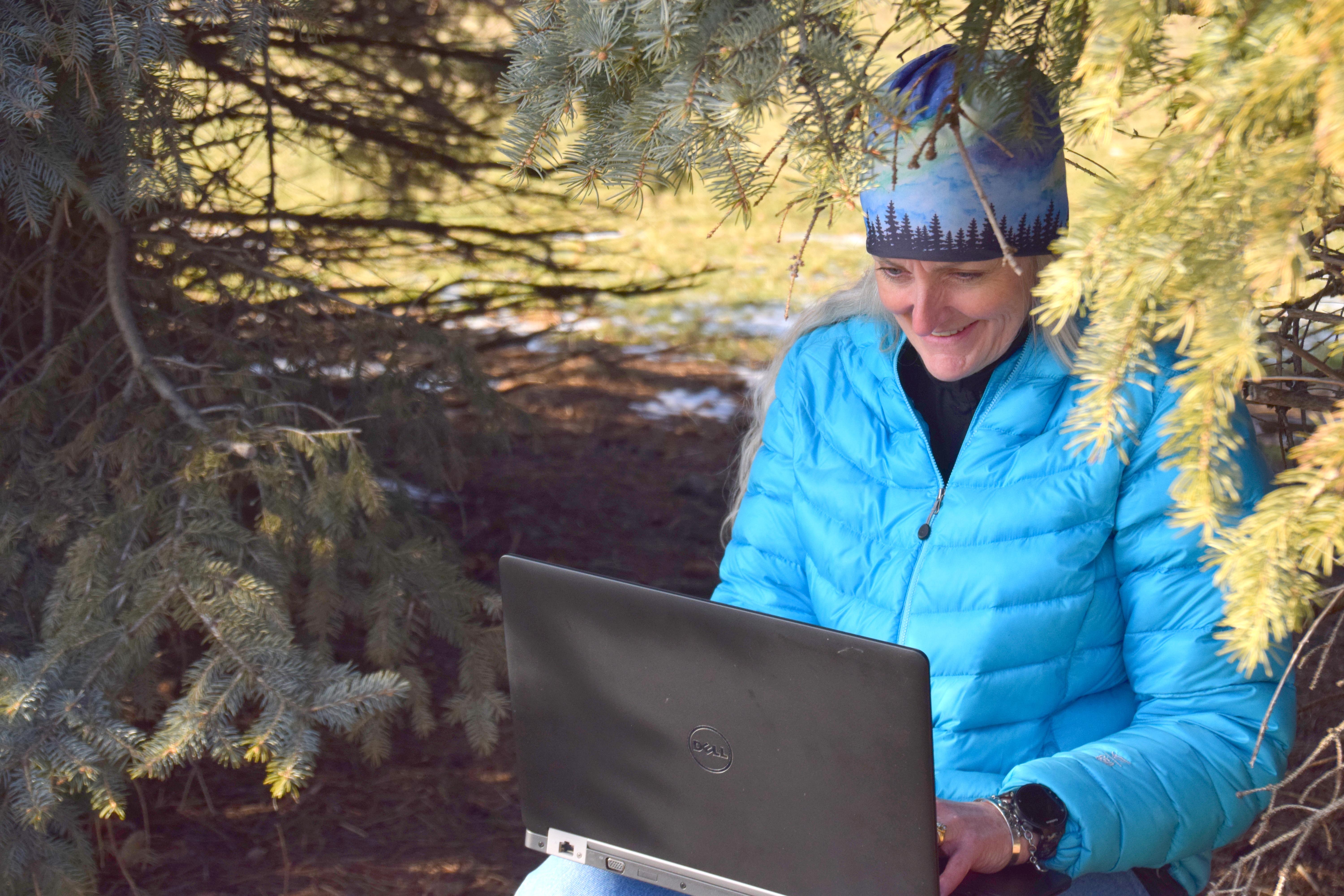 woman working on a laptop sitting under a pine tree