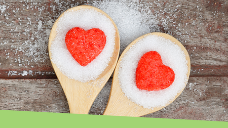 Two wooden spoons sitting on a wooden table. The spoons are full of white sugar with a red heart in the middle of each spoon.