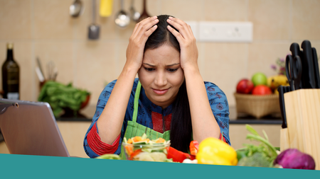 A stressed women with her elbows on the kitchen counter and her hands on top of her head while looking at different vegetables on the kitchen counter.