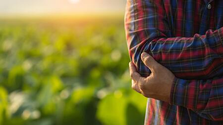 A Person standing near a farm field while holding their elbow as if it is sore.