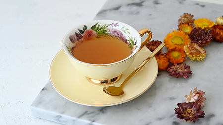 flowered tea cup on saucer with liquid in it an da golden spoon by dried flowers