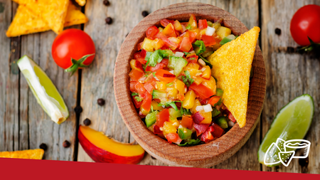 Apple mango salsa in a wooden bowl placed on a wood table with tomatoes, limes, and chips placed around the bowl on the table.