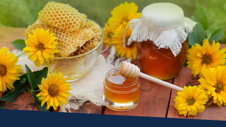 Honey comb and jars of honey sitting on a wooden table with daises surrounding the honey.