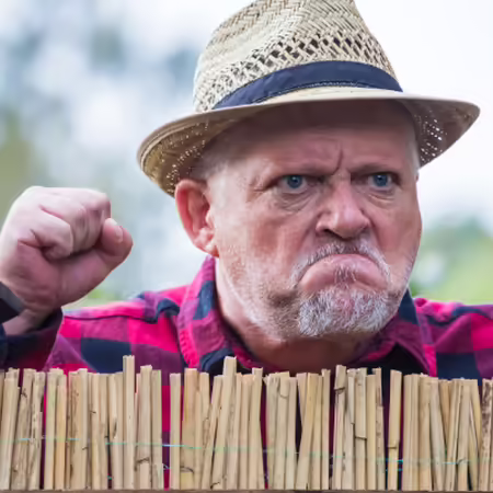 A man wearing a cap stands grimly behind a fence, glaring angrily at the neighbors.