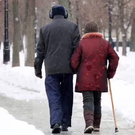 An older couple walking outside on a snowy sidewalk. One of them is using a walking cane.
