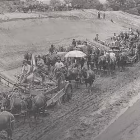 Historical black & white photo depicts large muddy farm field being dredged with equipment, many horses and people.