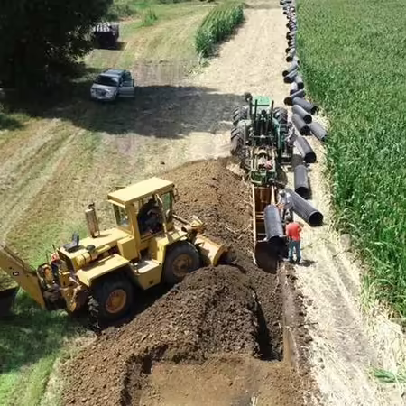 A tile contractor uses a backhoe loader to aid installation of a large tile main, with dirt road on left and farm field on the right.