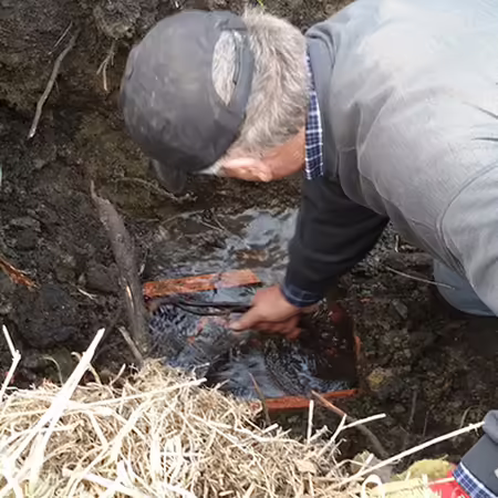 A farmer in cap and coveralls is bent over looking at a broken tile and water.
