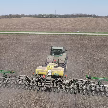 A large corn planter works in a field with blue sky and a road visible in the background.