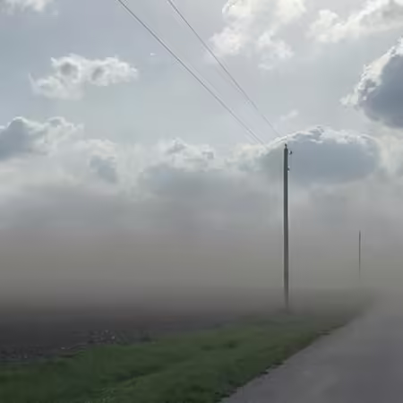 Sunshine peeks through large clouds over thick dust, which shrouds a country road running between two farm fields.