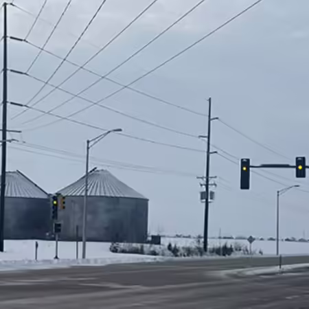 Two large grain bins on snow covered ground are visible at a stoplight intersection with a yellow light.
