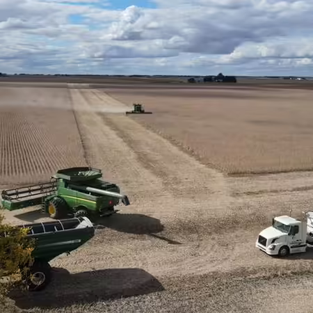 A large farming harvest operating is taking place in an agricultural field with clouds & blue sky in background.