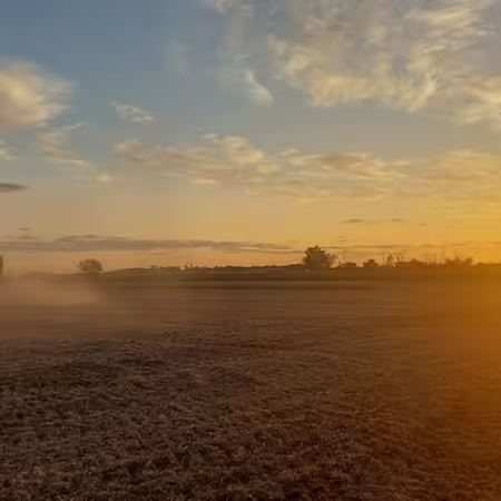 A combine moves across a dusty harvested field with clouds & setting sun in background.