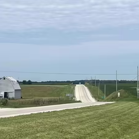 Distant view of a country road with a barn on the left, power lines on the right and a blue sky with some clouds in the background.
