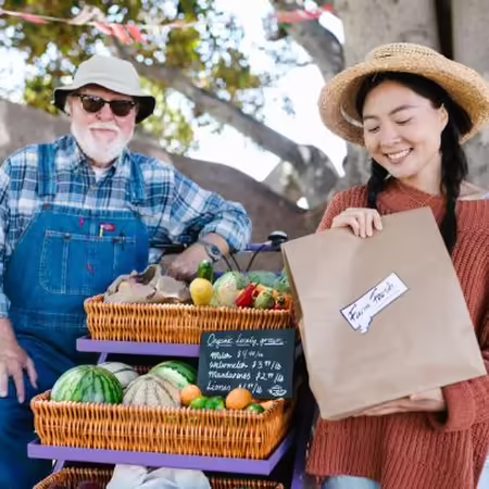 A woman in knitted sweater holding paper bag with a famer both standing next to woven basket filled with fruits