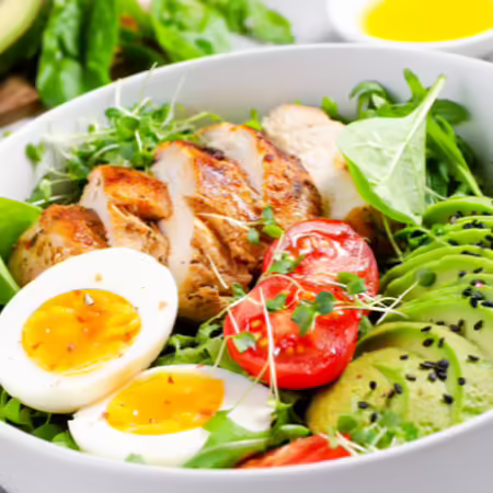 White bowl with salad, salad containing boiled egg, chicken, tomatoes, avocado and salad greens. 