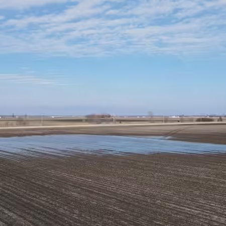 Photo shows wide shot of unplanted farm field with the far 2-3 foot edge flooded with rainwater. On the horizon a few trees, blue sky and some billowy clouds are visible.