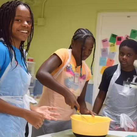 children with aprons, one is stirring food in a bowl.