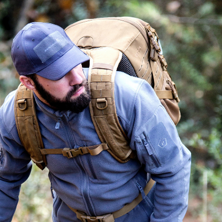 White man with dark beard in blue shirt and hat hiking with backpack