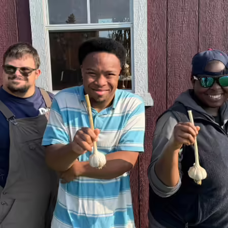 Two Rooted for Good employees stand next to 3 people proudly showing off their garlic harvest