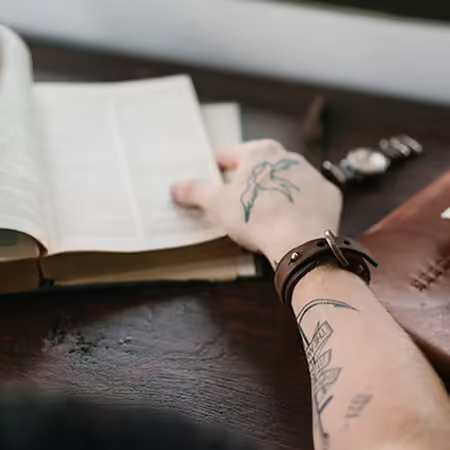Man with tattoos reading a book with wallet nearby