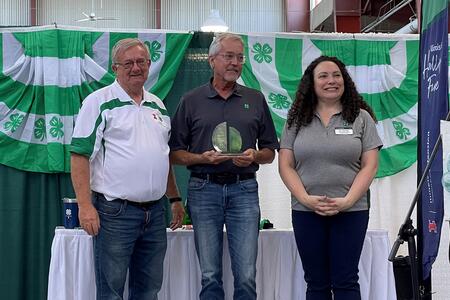 3 people on a stage in front of 4-H bunting; center person holding an award