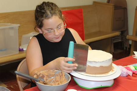 Girl in black shirt putting frosting on a round cake.