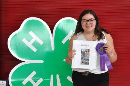 4-H member holding project and ribbon in front of large 4-H Clover backdrop