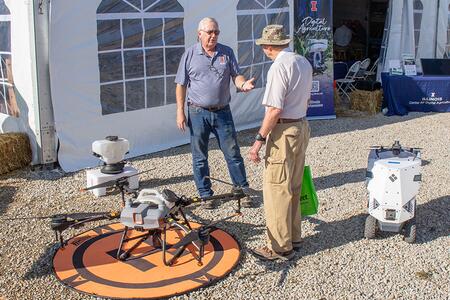 Two people talking about agriculture with a spray drone and robot seeder nearby.
