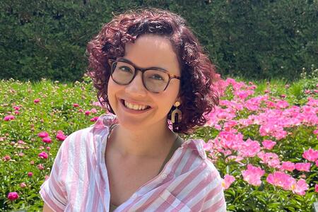 A woman with glasses and curly hair outside in front of pink flowers