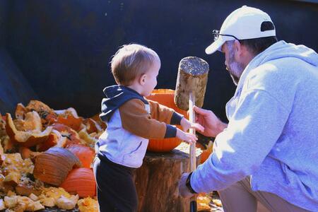 An adult assists a young child with smashing a pumpkin at a composting event.