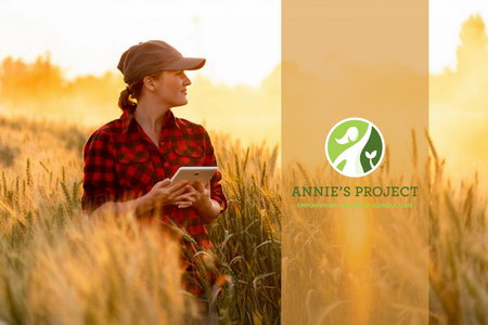 a woman in a red and black shirt and brown baseball cap standing in a field of crops with sunlight at her back. She is looking to her left across the field and holding a tablet.