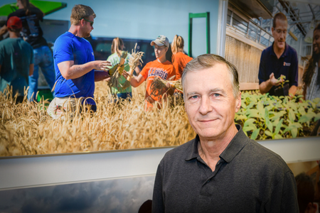 A man, Lowell Gentry, in front of a photo of a farm field