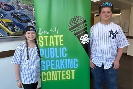 2 youth wearing baseball jerseys standing in front of a banner that says "Illinois 4-H State Public Speaking Contest"