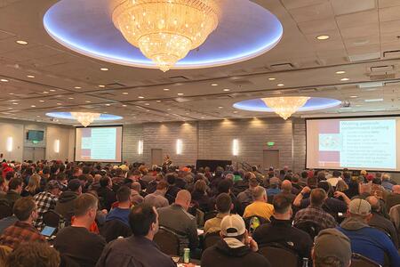 A large room completely filled with people sitting in chairs at tables listening to a training presentation.