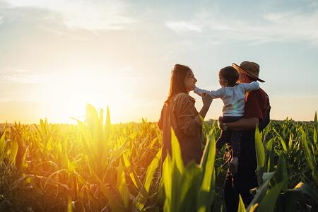 A family of three stand in a farm field overlooking a sunset