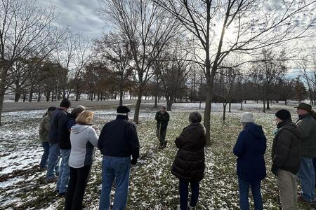 A man leans on a tool outside by a tree surrounded by a group of people