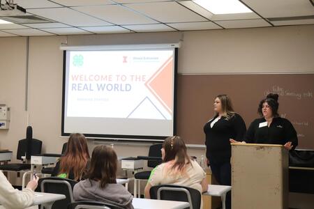Two women standing at a lectern with the presentation slide that says, "Welcome to the Real World".