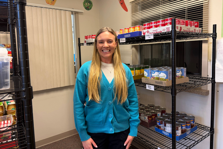 Morgan is pictured in a blue jacket, standing in front of a food pantry display