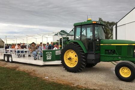 People riding on a seated trailer being pulled around a field day event by a green tractor.